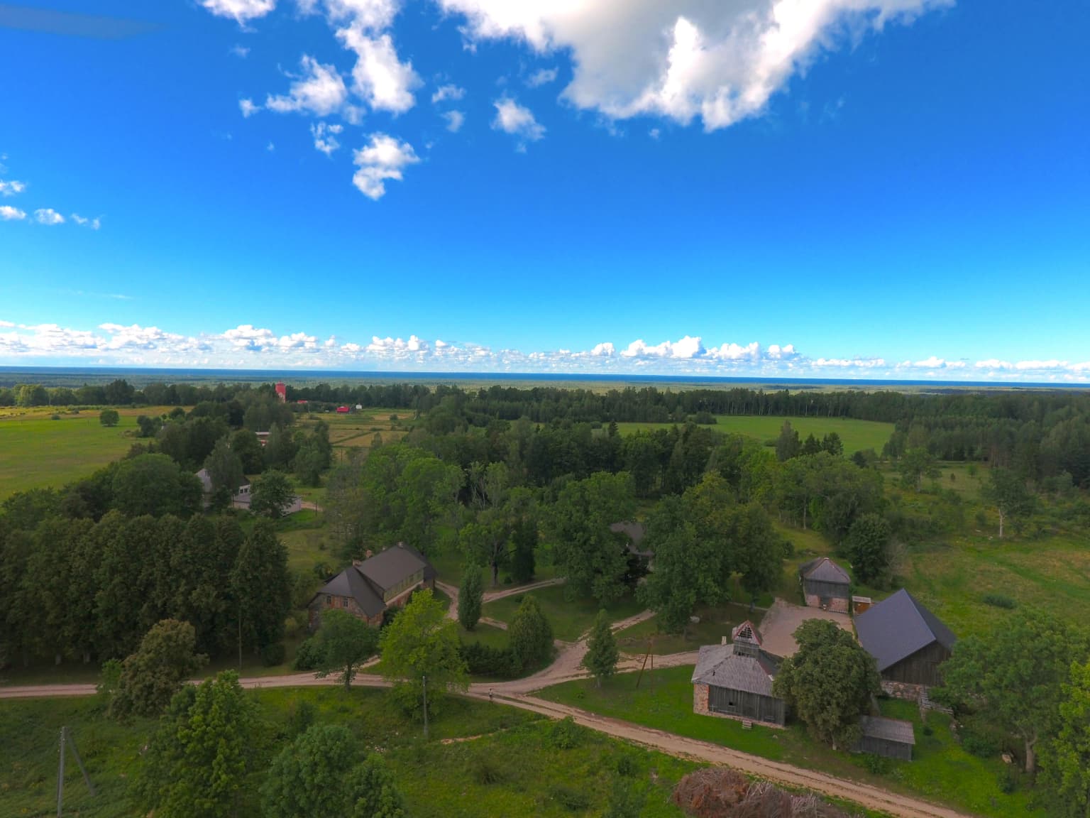 Aerial view of a landscape with green fields, dense forests, several houses, and dirt roads under a bright blue sky with scattered clouds