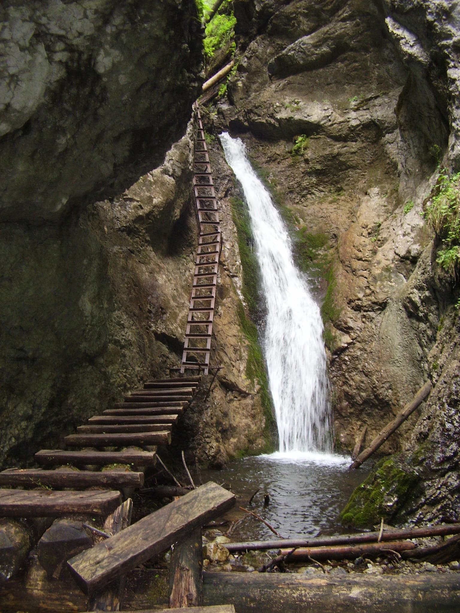 Waterfall flowing down rocky cliffs into a pool with wooden ladder structure on left side