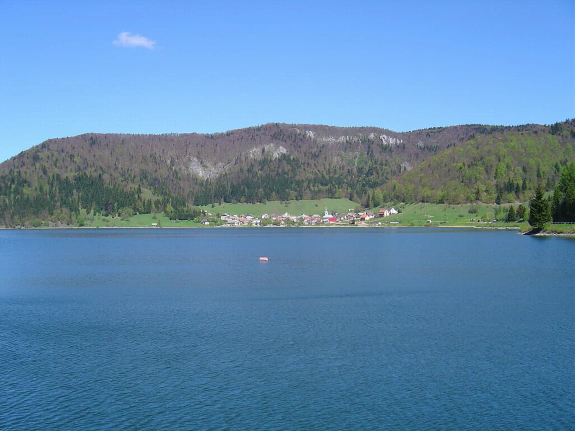 Large lake with small boat, surrounded by forested hills and village under clear blue sky