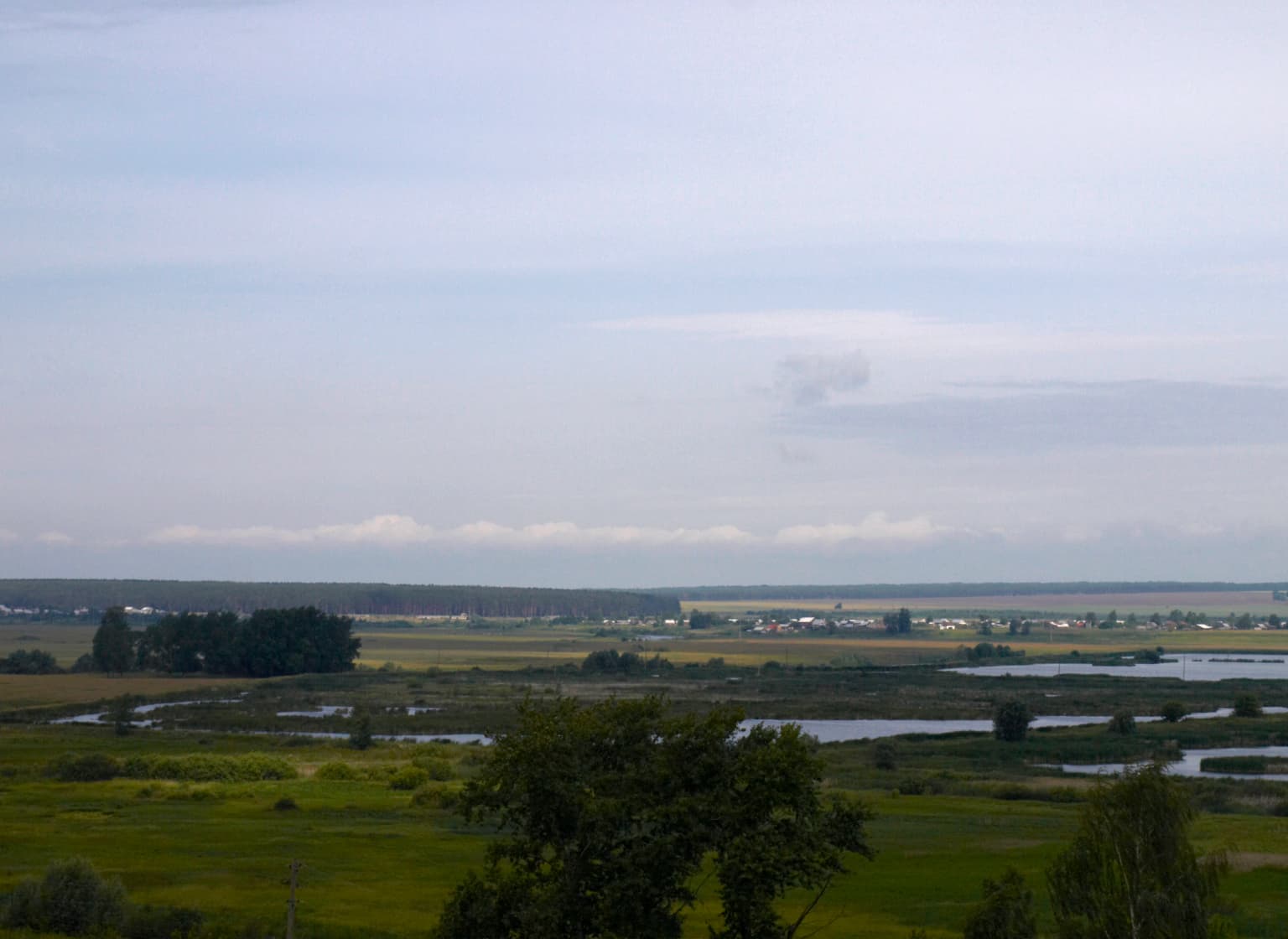 Wide landscape view showing the Alatyr River flowing through wetlands with fields, trees, and distant settlements under a partly cloudy sky