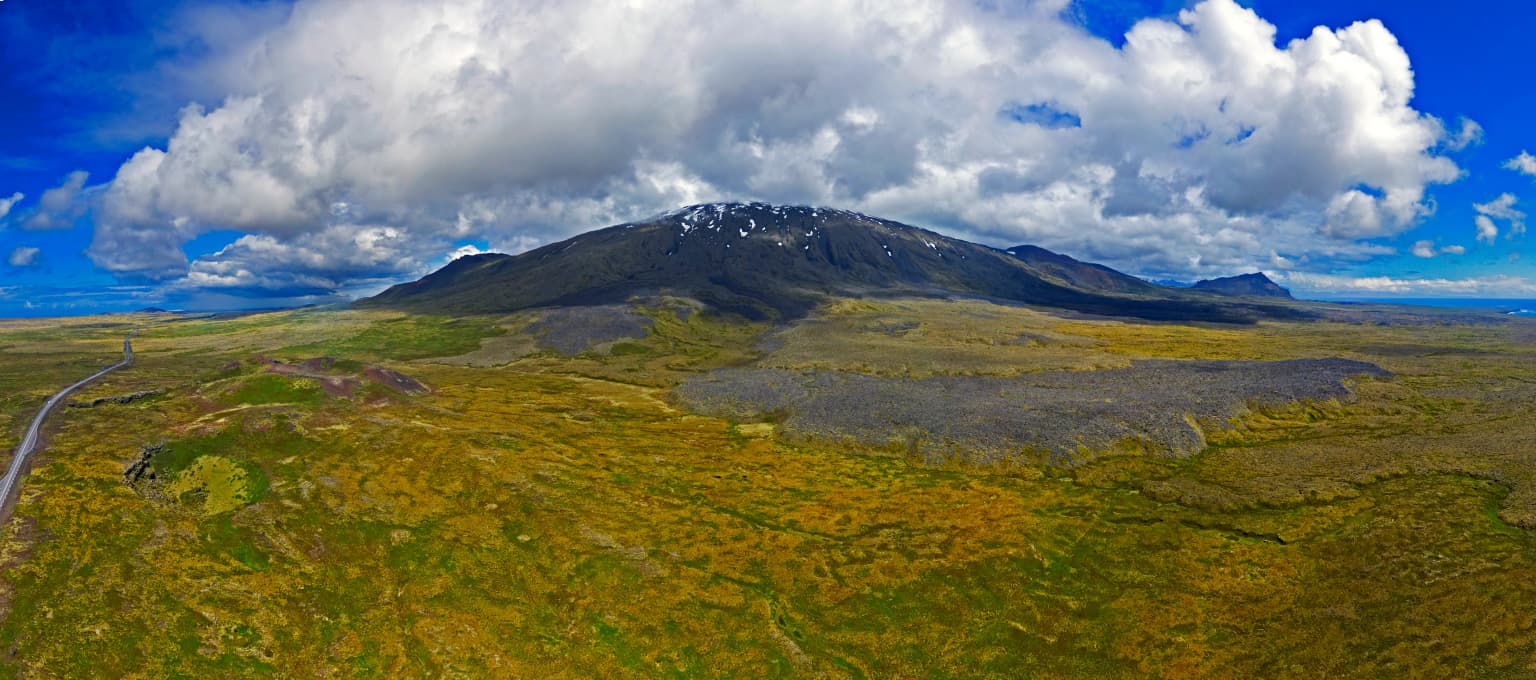 Aerial view of Snæfellsjökull volcano showing snow-covered peak, rolling green and yellow fields, a winding road, and scattered clouds against a blue sky