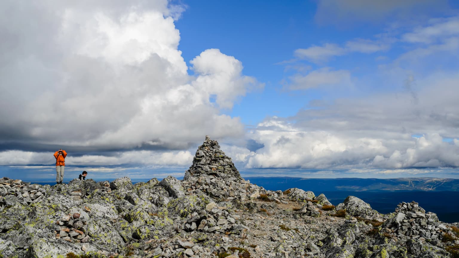 Rocky mountain summit featuring a stone cairn, two small figures of people, and a cloudy sky