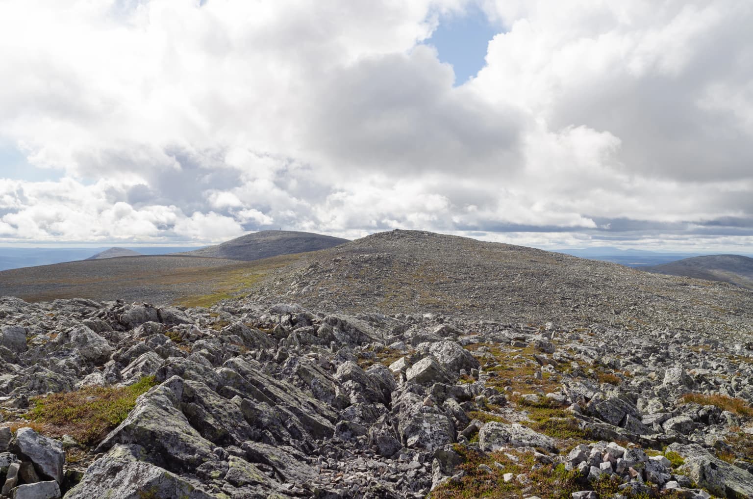 Rocky terrain with patches of moss and lichen in the foreground, extending to a flat-topped mountain summit under a partly cloudy sky with distant mountains visible on the horizon