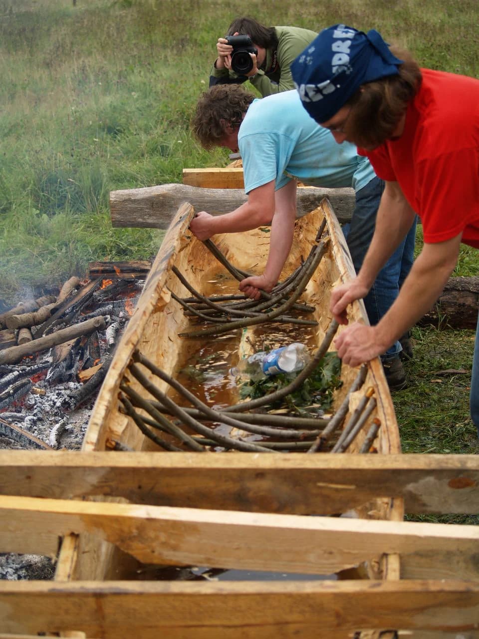 Three people working on a wooden dugout canoe with a fire and grassy background