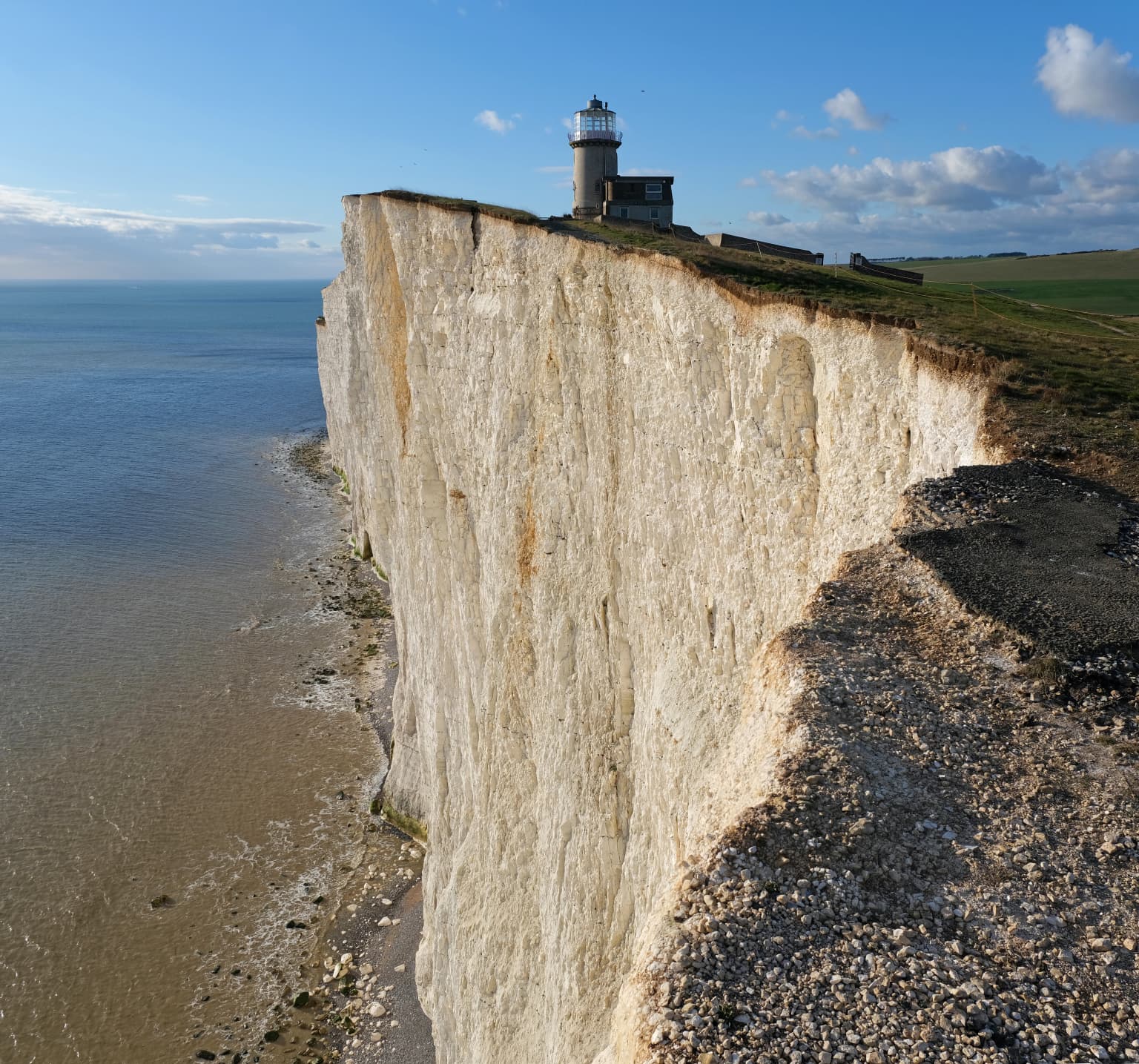 White chalk cliffs with a lighthouse on the cliff edge overlooking the sea and rocky beach.