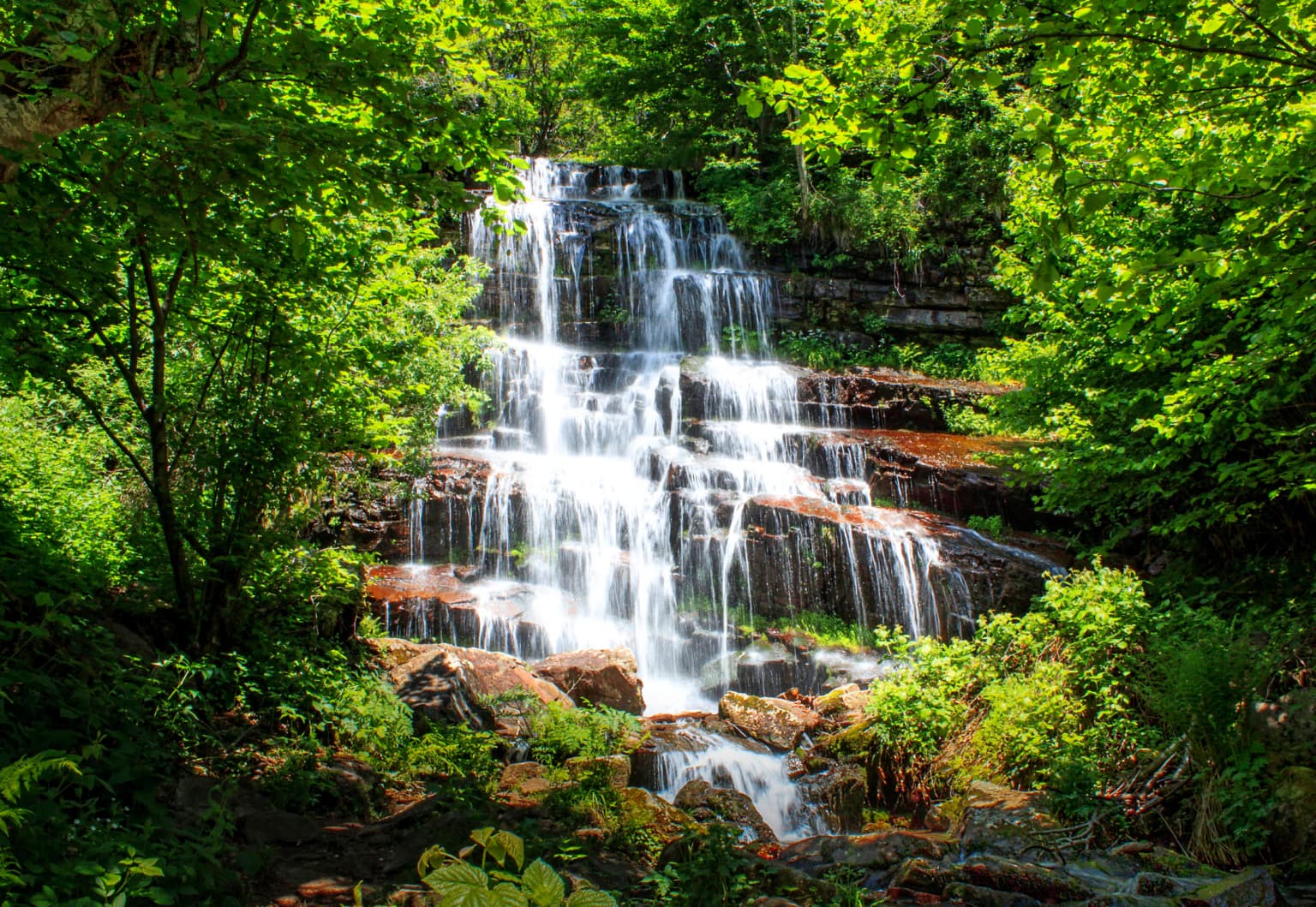 A multi-tiered waterfall cascading over rocks surrounded by lush green trees and vegetation
