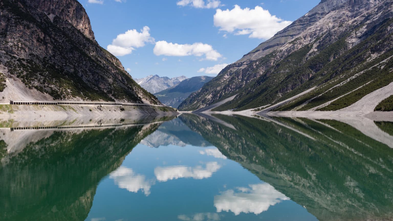 Calm lake reflecting steep rocky mountains and white clouds under a clear blue sky