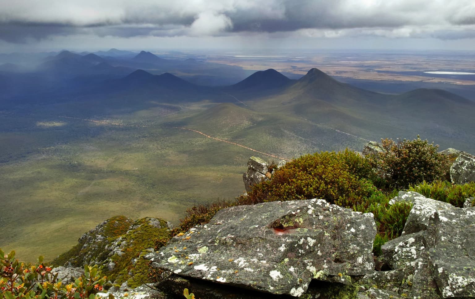 Vista panorámica del paisaje desde una cumbre montañosa que muestra terreno rocoso con líquenes, vegetación baja y colinas onduladas distantes bajo un cielo nublado