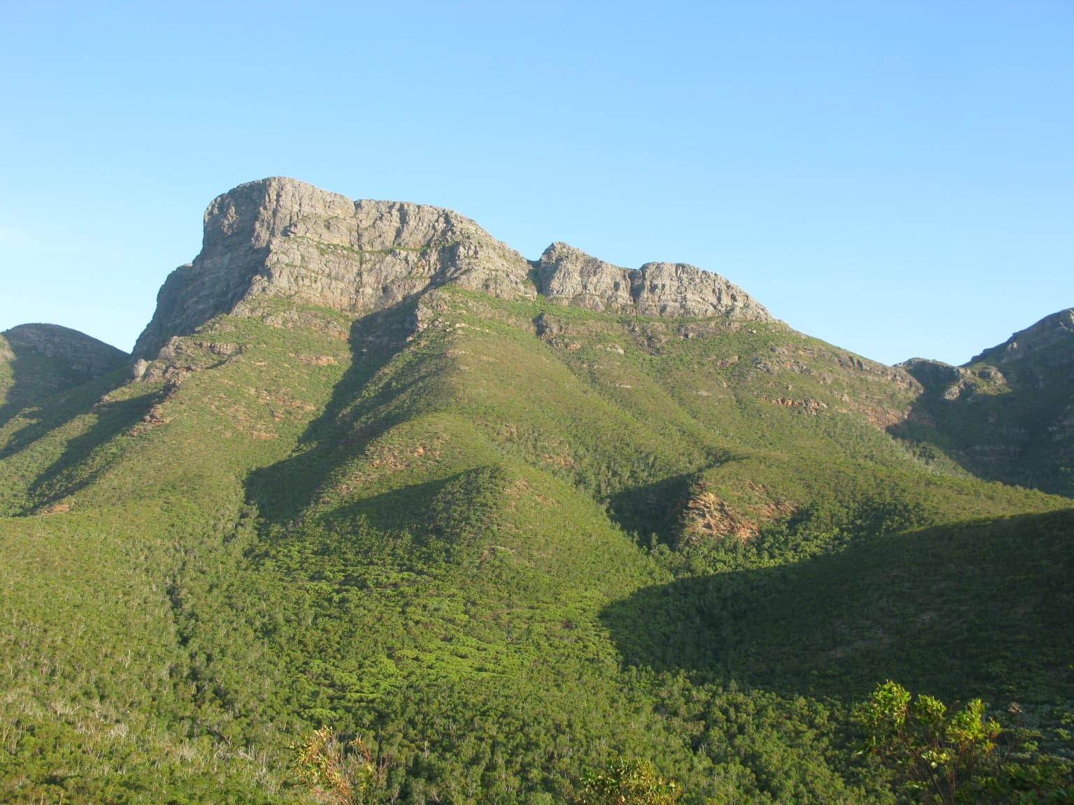 Una cumbre montañosa con cima rocosa y laderas exuberantes y verdes bajo un cielo azul despejado.