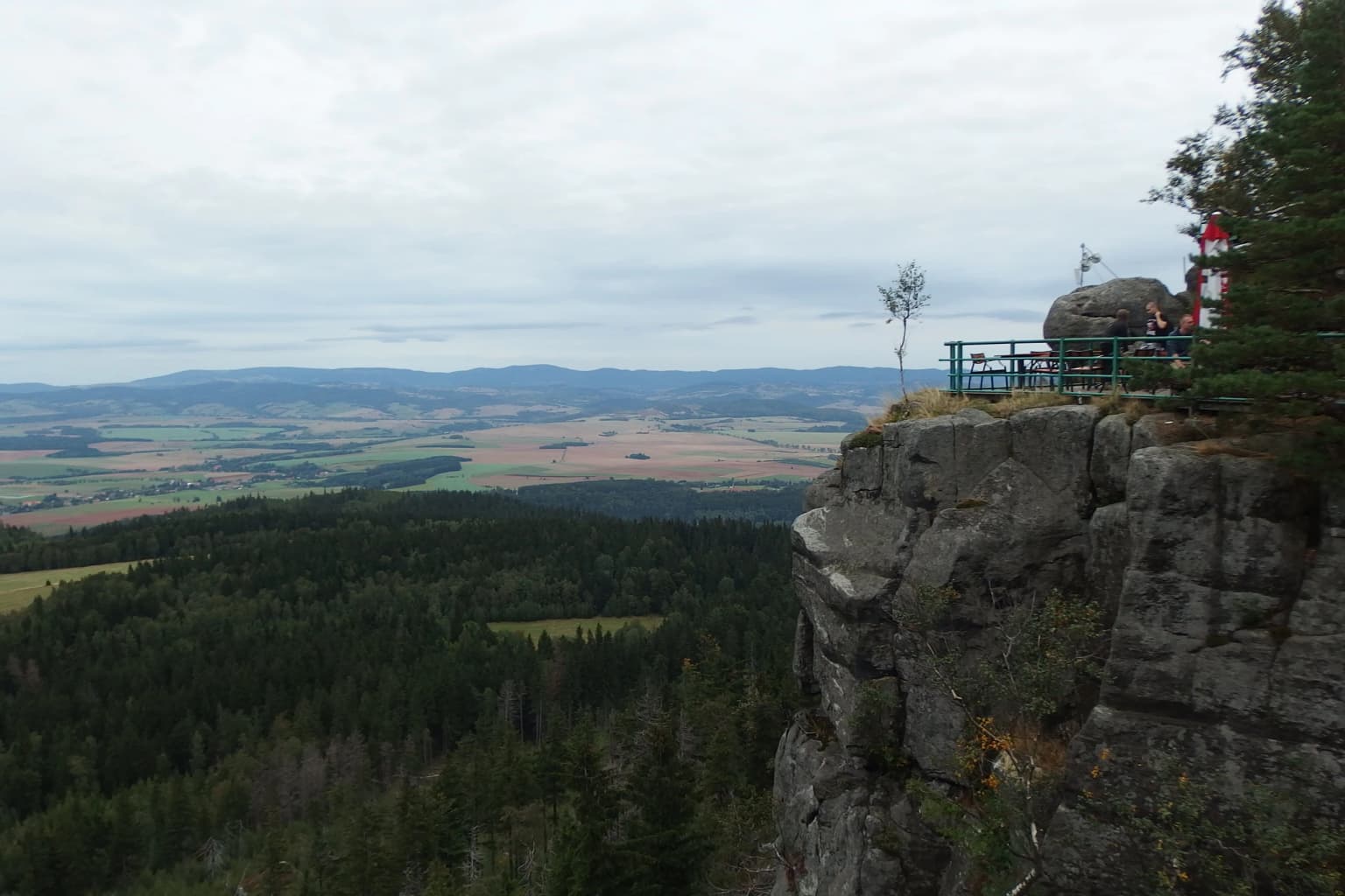 Rocky sandstone cliff edge with metal railing and visitors, overlooking dense forested valleys and distant mountains under overcast sky