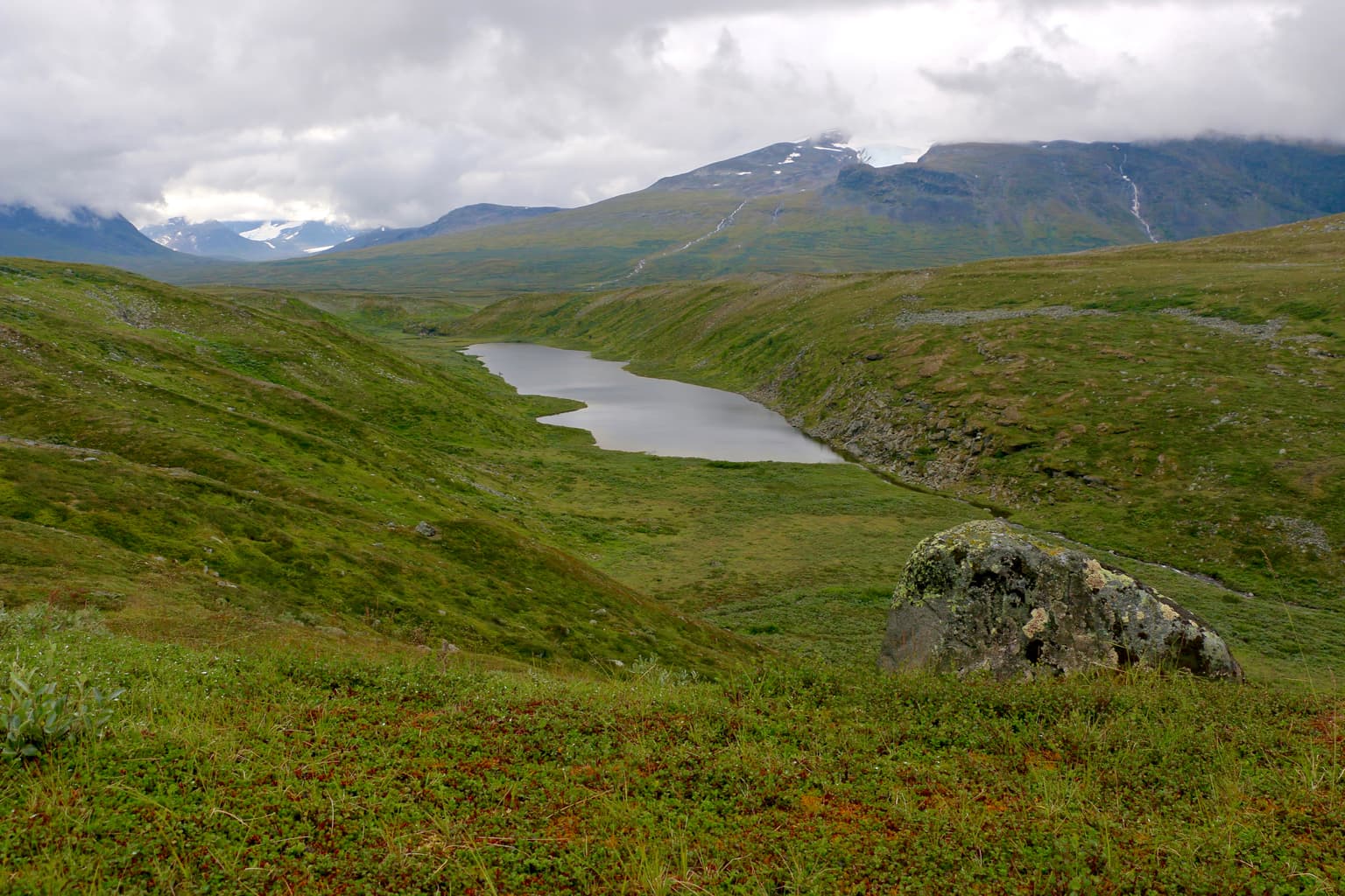 A wide landscape view of a lake surrounded by rolling green hills and mountains under a cloudy sky