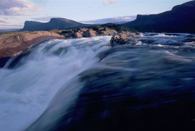 Waterfall flowing over rocks with mountains visible in the distance under a partly cloudy sky