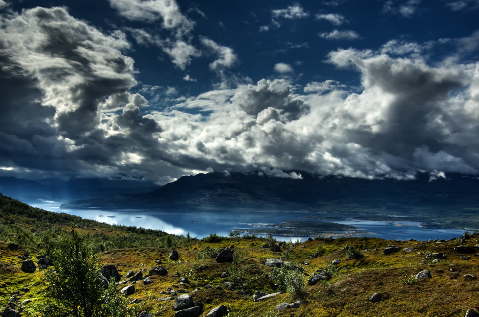 Mountainous landscape with a lake under dramatic clouds, showing open terrain with rocks and sparse vegetation
