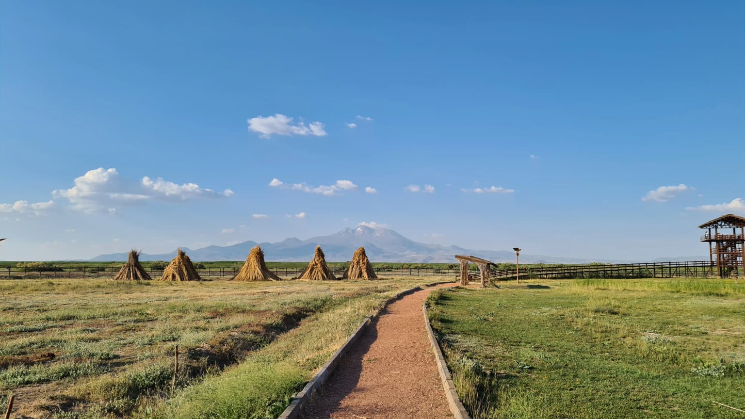 Dirt path through grassy field with hay stacks, wooden structure, and Mount Erciyes in the distance under a clear blue sky