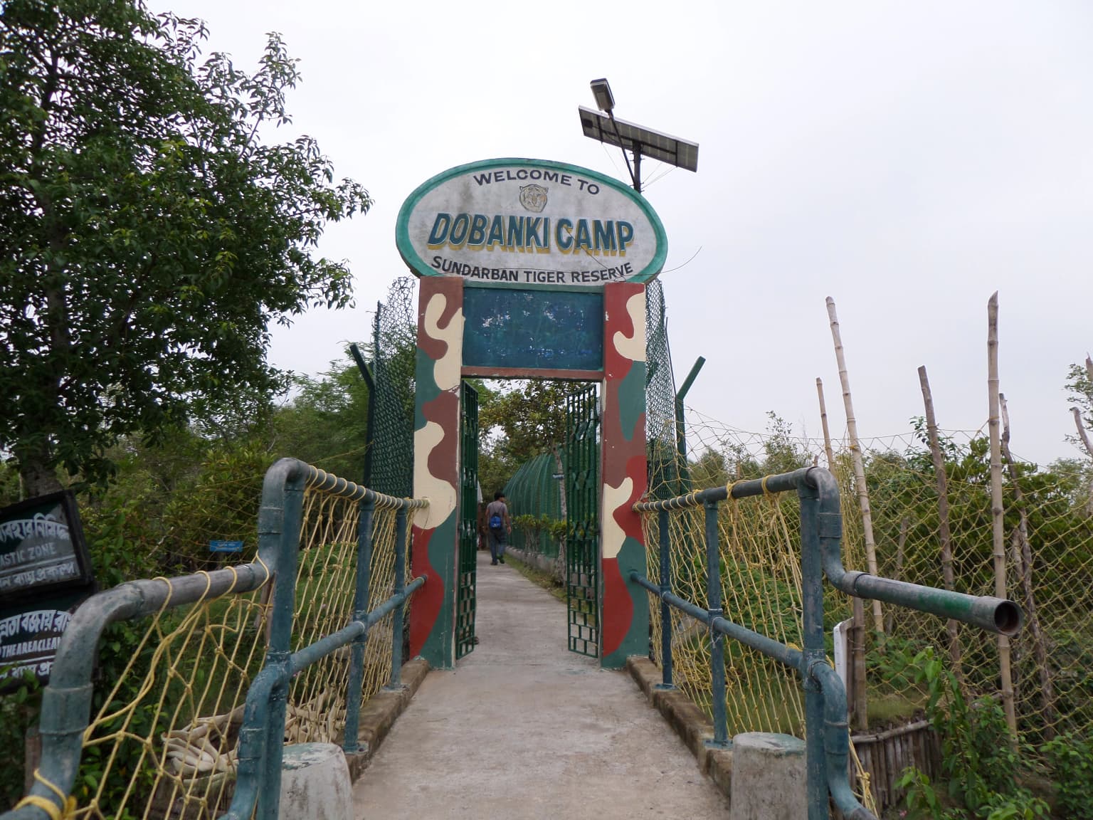 Estructura de valla metálica verde con un cartel de 'Welcome to Dobanki Camp' sobre un sendero, rodeada de vegetación, con una persona caminando al fondo