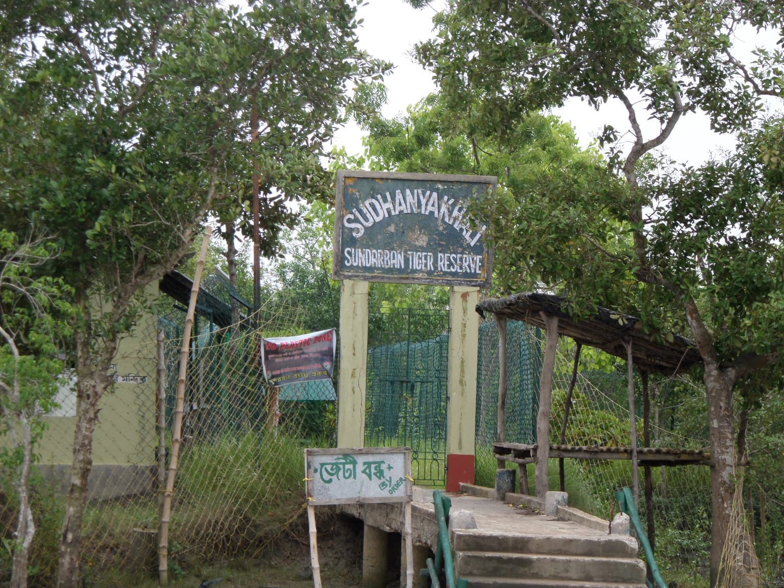 Entrance sign for Sudhanyakhali area of Sundarbans Tiger Reserve with Bengali text below, surrounded by trees and vegetation