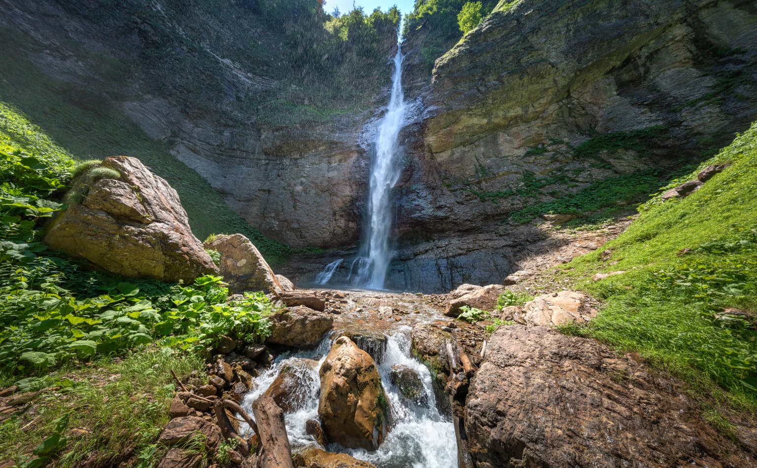 Waterfall flowing down a rocky cliff with green vegetation on both sides and a stream below