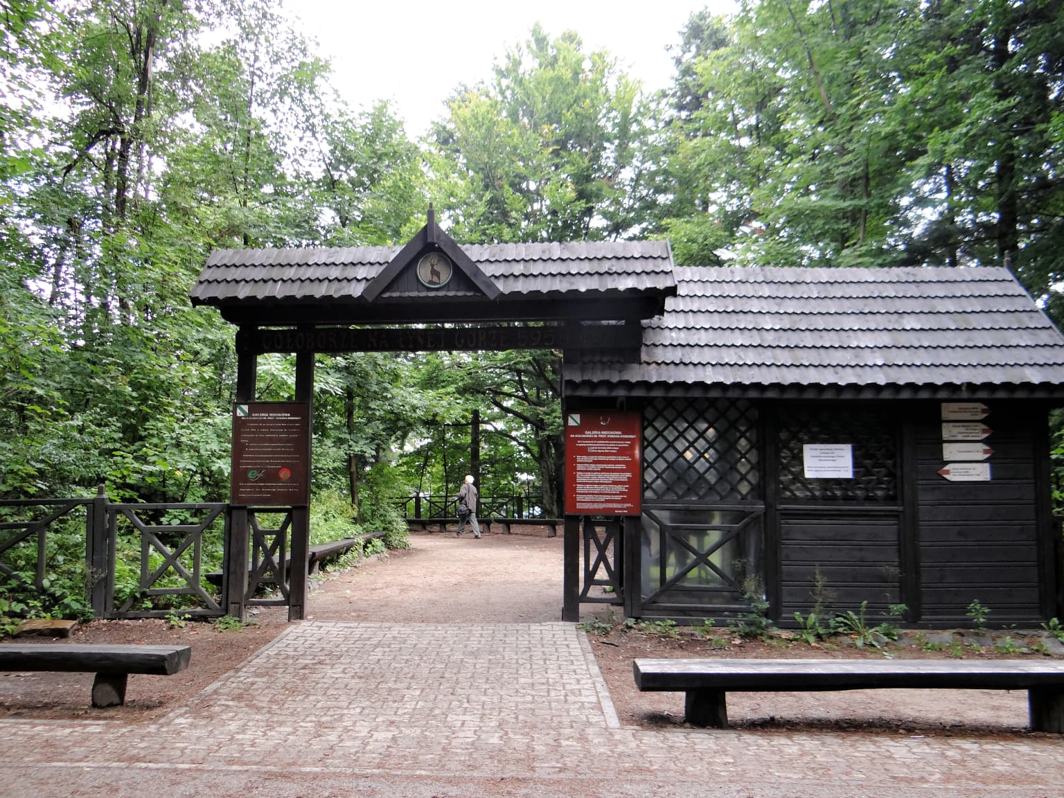 Wooden entrance gate with roof, adjacent building, benches, paved path, and forest background
