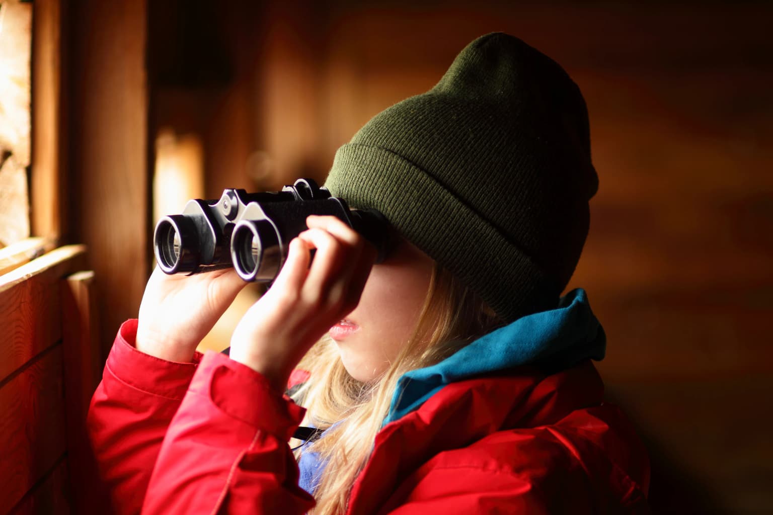 A girl in a red jacket and green beanie using binoculars to look out of a wooden shelter window