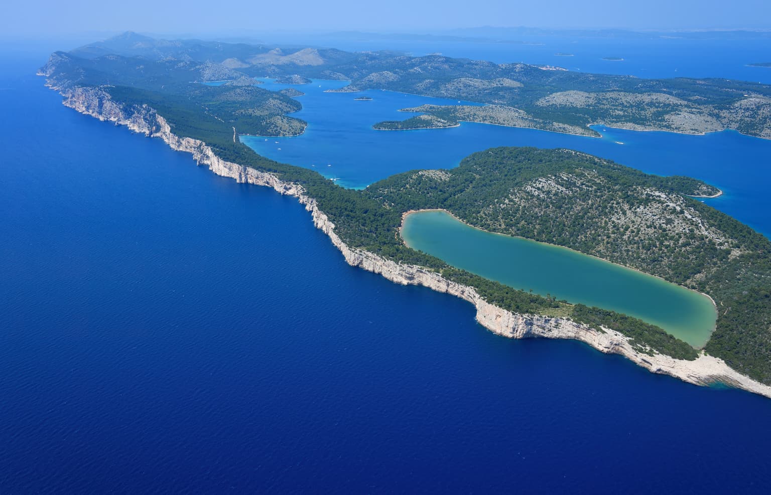 Aerial view of Telašćica Nature Park showing blue sea, rocky coastline, and turquoise saltwater lake surrounded by green terrain