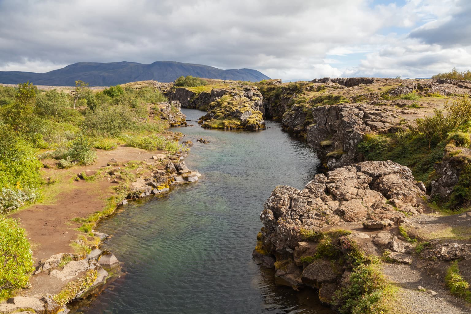 A river flows through a rocky canyon with green vegetation on the banks and mountains in the background