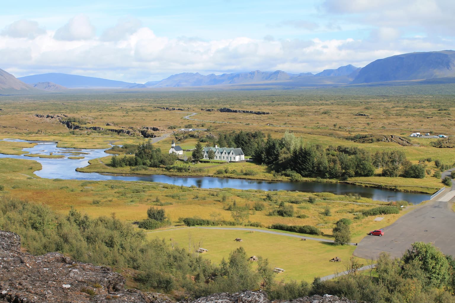 Wide landscape view showing a river winding through grassy fields with a small building, trees, and mountains in the background under a partly cloudy sky.