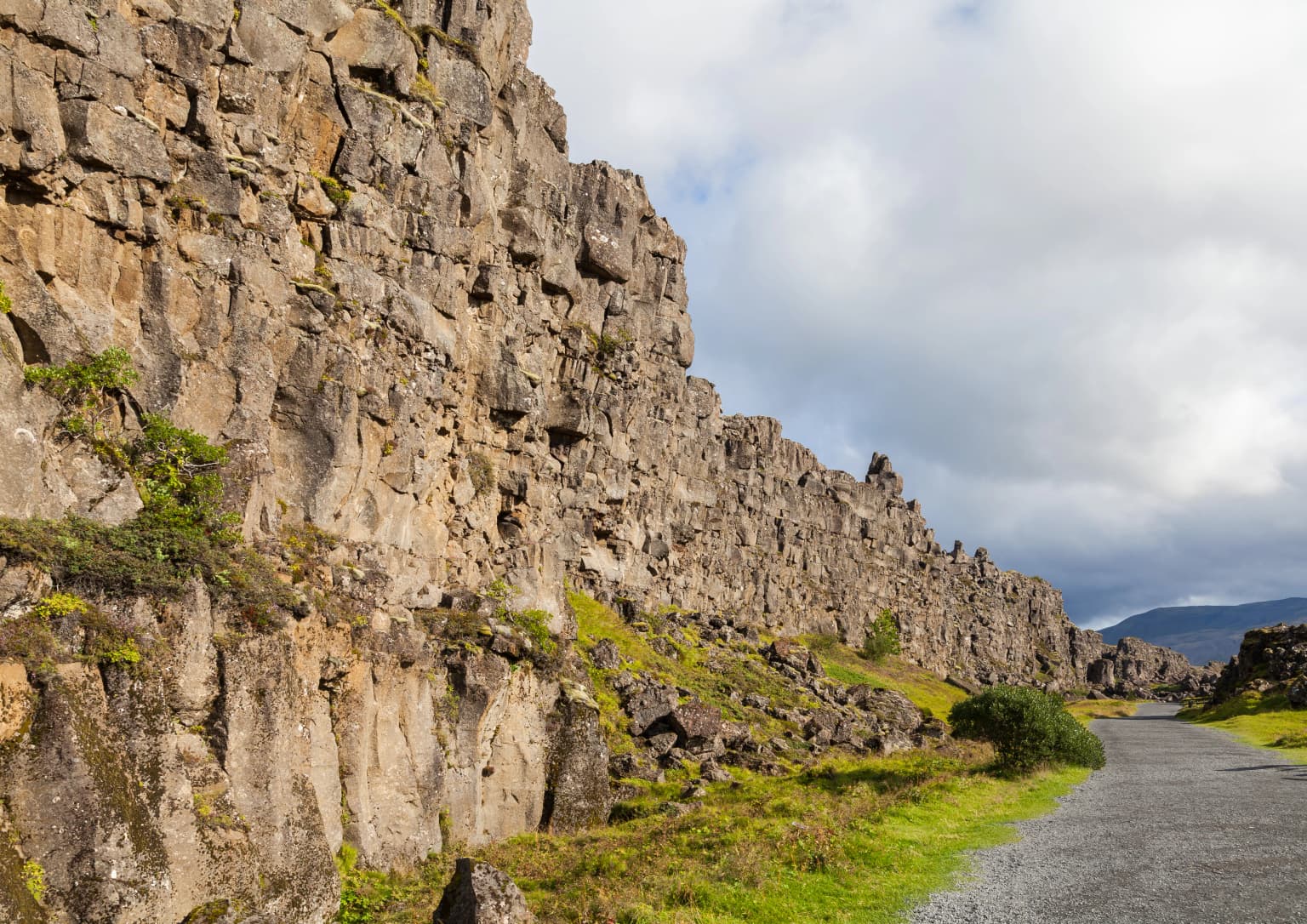 Tall rocky cliff face with patches of green vegetation alongside a gravel road under partly cloudy sky