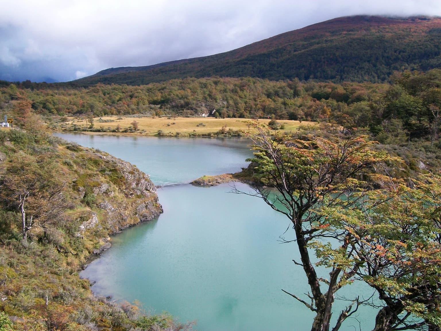 A turquoise river winds through a landscape of rocky hills and autumn-colored trees, with forested mountains in the background under a cloudy sky