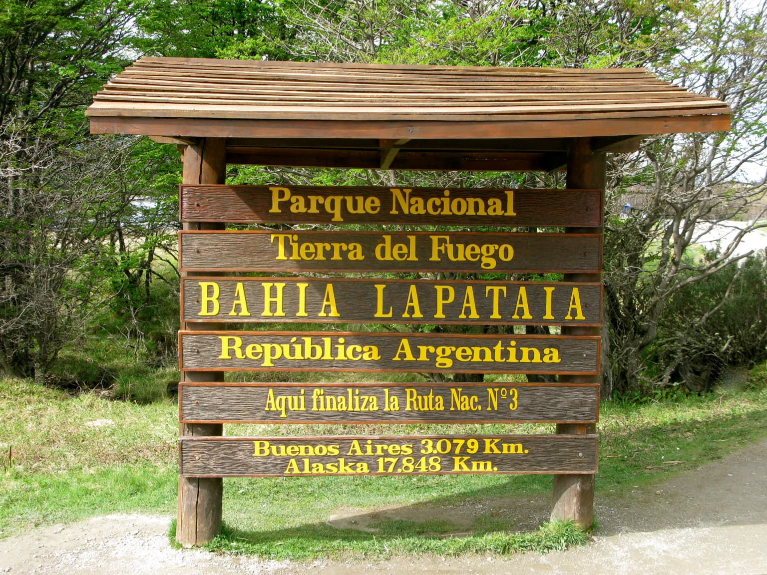 Wooden sign structure with multiple plaques displaying 'Parque Nacional Tierra del Fuego', 'Bahia Lapataia', 'República Argentina', 'Aqui finaliza la Ruta Nac. N°3', 'Buenos Aires 3079 Km', 'Alaska 17348 Km' in yellow text on brown planks, surrounded by green grass and trees