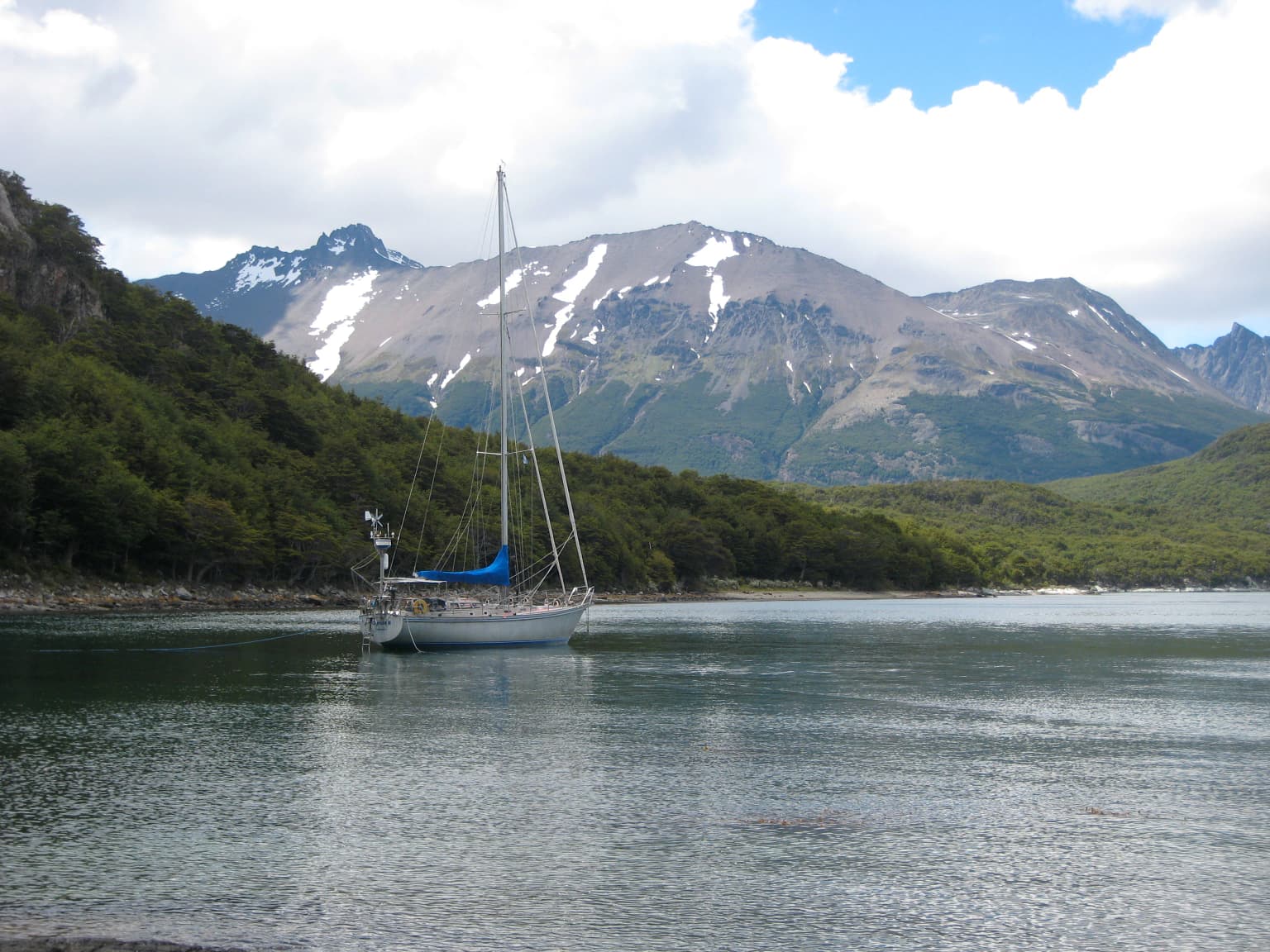 Velero flotando sobre aguas tranquilas con colinas boscosas y montañas nevadas de fondo bajo un cielo parcialmente nublado