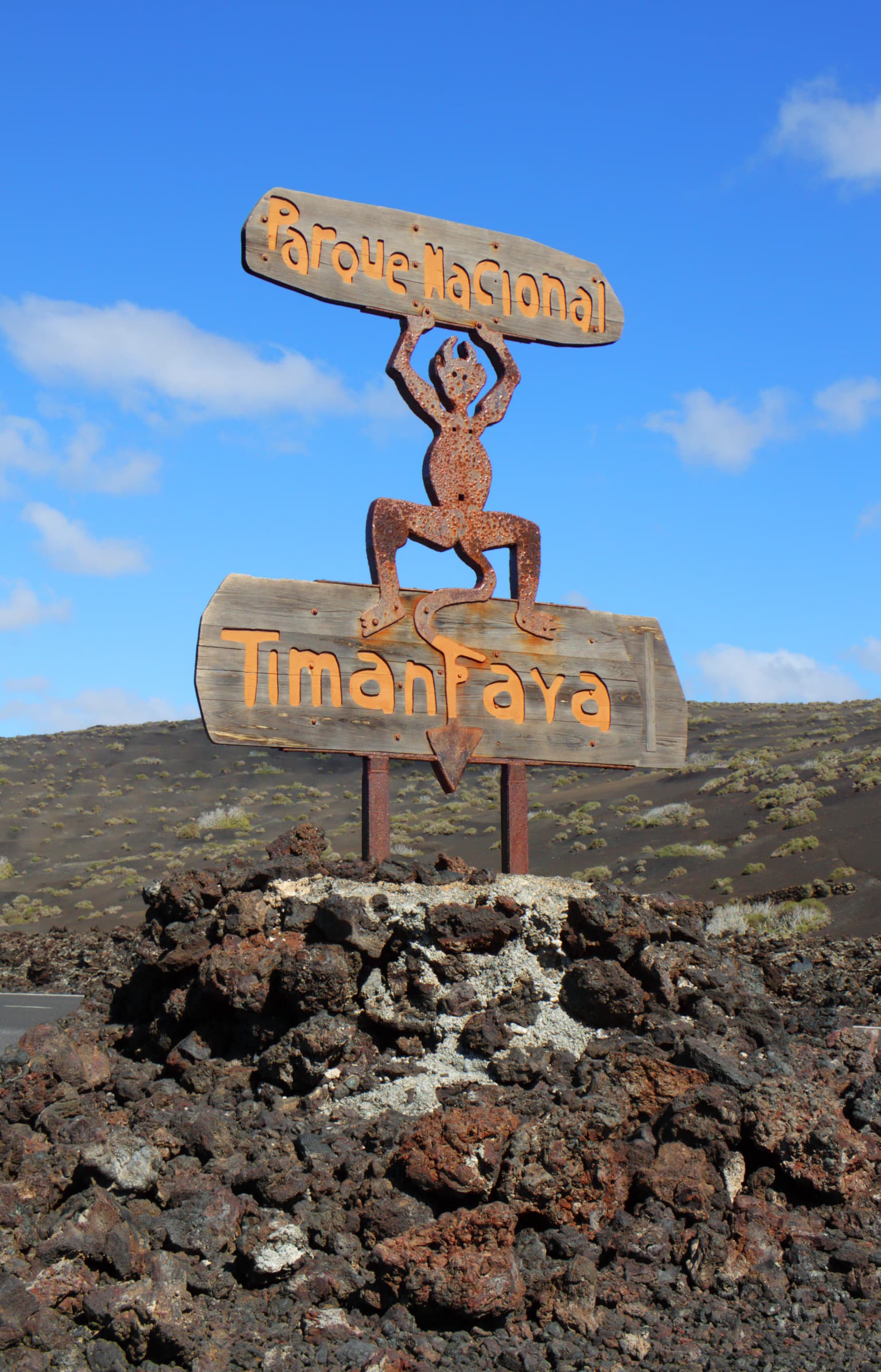 Wooden entrance sign for Timanfaya National Park with 'Parque Nacional' and 'Timanfaya' text, supported by a rusted metal sculpture of a figure holding the top sign, set against rocky volcanic terrain under a clear blue sky.