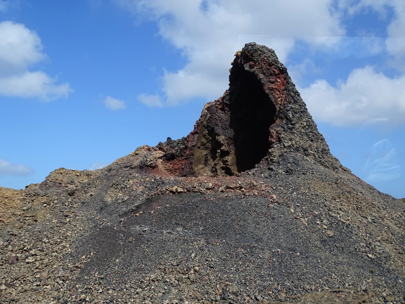 A rocky volcanic cone with a dark crater opening under a blue sky with scattered clouds