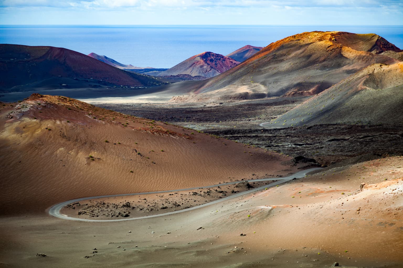 Wide scenic view of Timanfaya National Park showing volcanic hills with earth-toned slopes, a winding road through barren terrain, and distant mountains under a clear sky