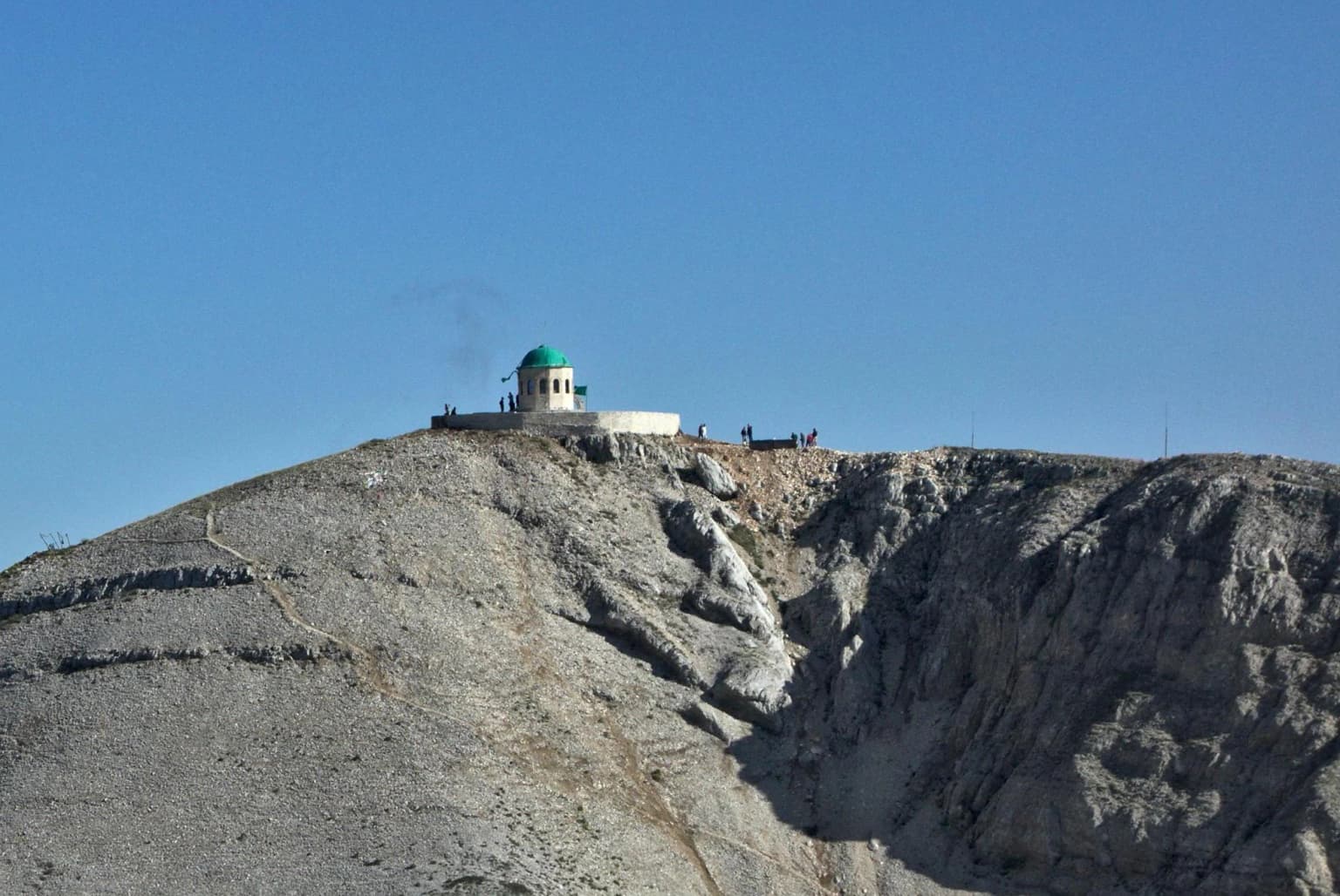 Rocky mountain peak with white shrine featuring green dome, small groups of people visible, clear blue sky