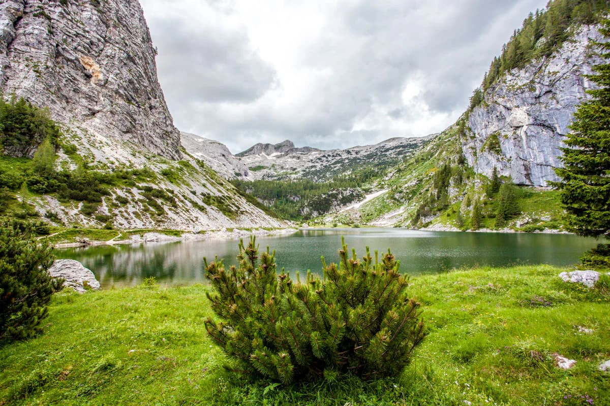 A clear lake surrounded by steep rocky cliffs and lush green meadows under a partly cloudy sky