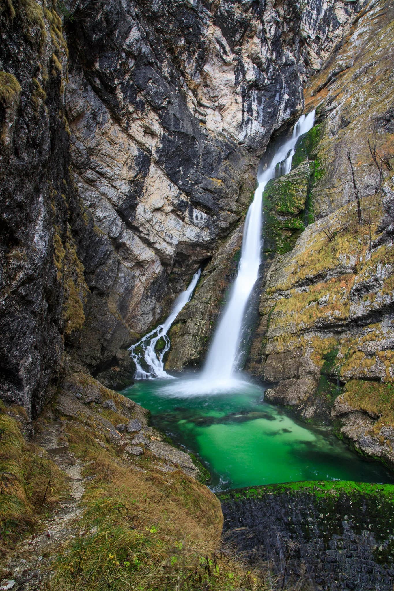 Waterfall flowing into a greenish pool between steep rocky cliffs with a narrow trail on the left
