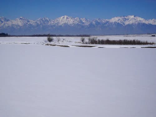 Snow-covered field with the Irkut River and distant snow-capped mountains under a clear blue sky