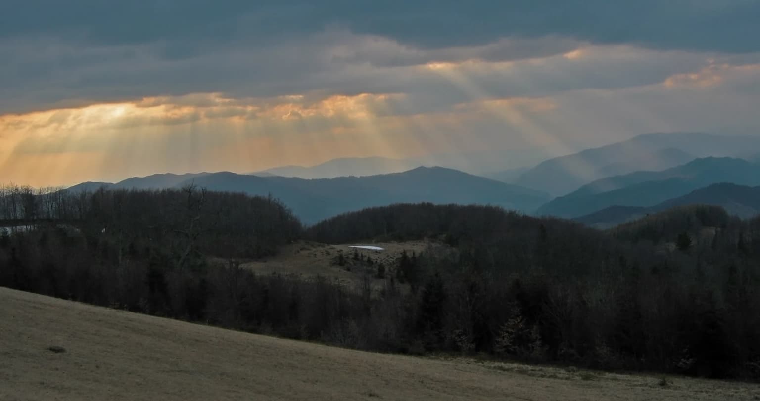 Sunlight rays breaking through clouds over a mountainous landscape with forested hills and open fields.