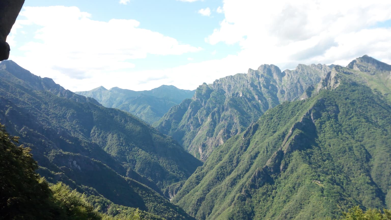 Panoramic mountain landscape with green valleys, rocky peaks, and scattered clouds under a bright sky