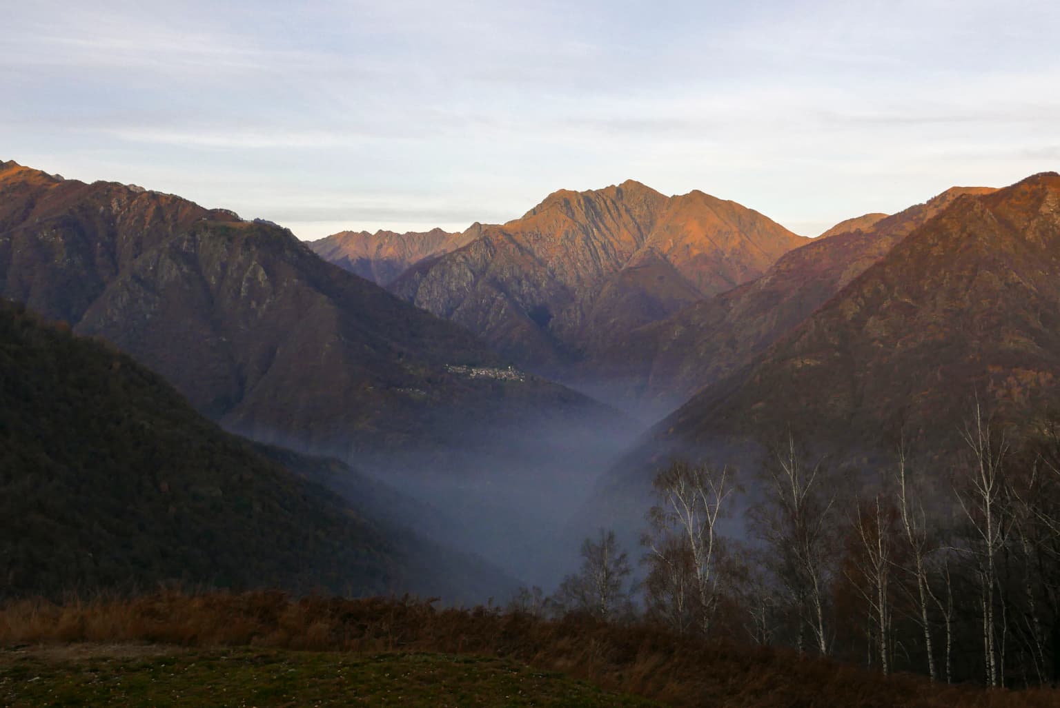 Wide view of mountain valley with mist, forested slopes, and distant ridges under a partly cloudy sky