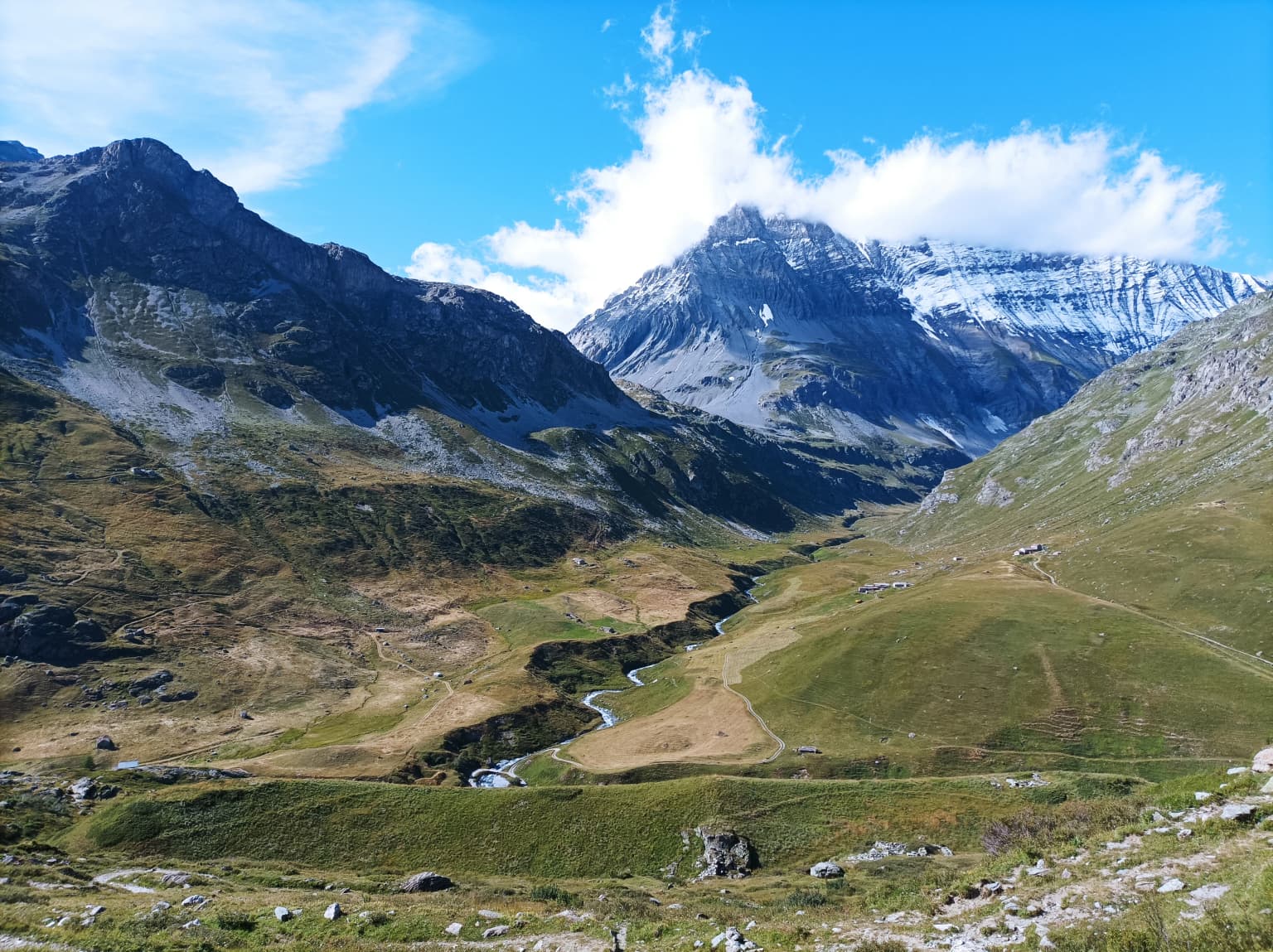 Wide landscape view of a mountain valley with Grande Casse peak in the background, green slopes, winding river, and scattered rocks under a blue sky with clouds.