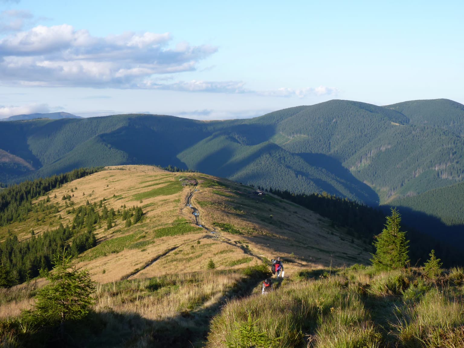 A scenic road ascending a grassy hillside with rolling mountains and forests under a partly cloudy sky.