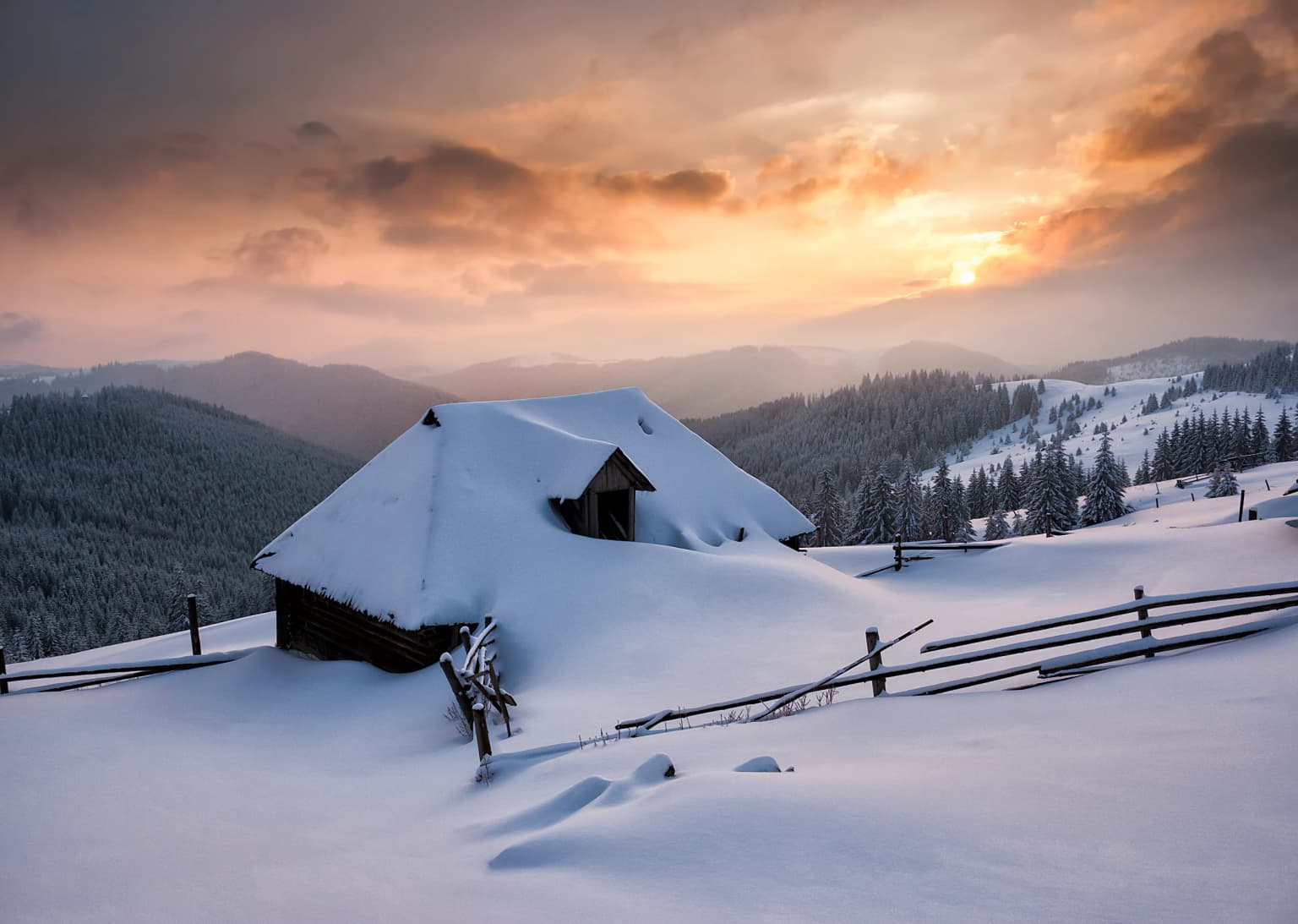 Snow-covered wooden cabin with a sloped roof, surrounded by a snow-covered landscape, wooden fence, and mountainous terrain under a partly cloudy sky with sunset hues