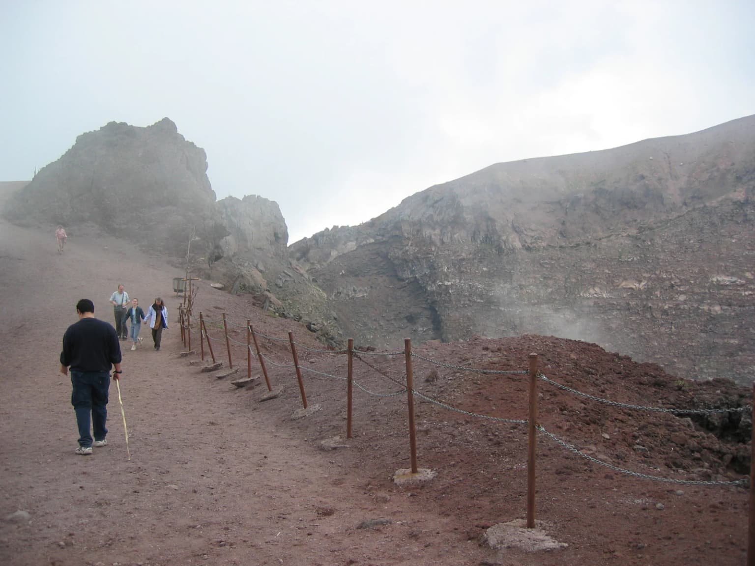 Hikers walking on a dirt trail with a roped barrier system, approaching a mountainous landscape with rocky terrain and mist