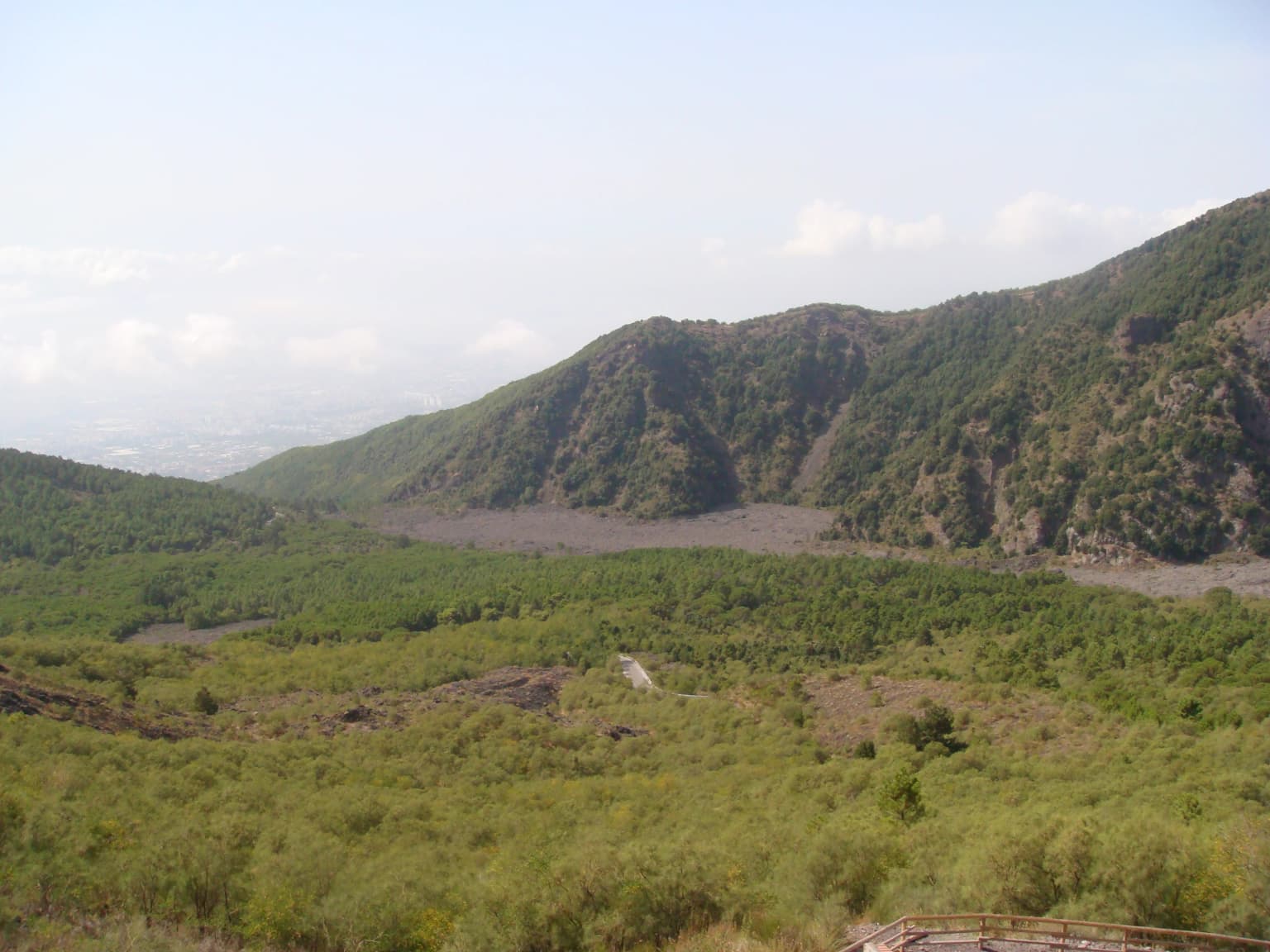 Wide landscape view of the valley between Monte Somma and Vesuvius mountains with green vegetation and forested hills under a partly cloudy sky