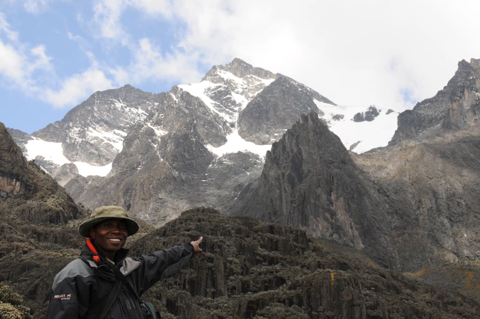 Una persona con gorra verde y chaqueta oscura se encuentra delante de montañas escarpadas y nevadas con terreno rocoso, bajo un cielo parcialmente nublado.