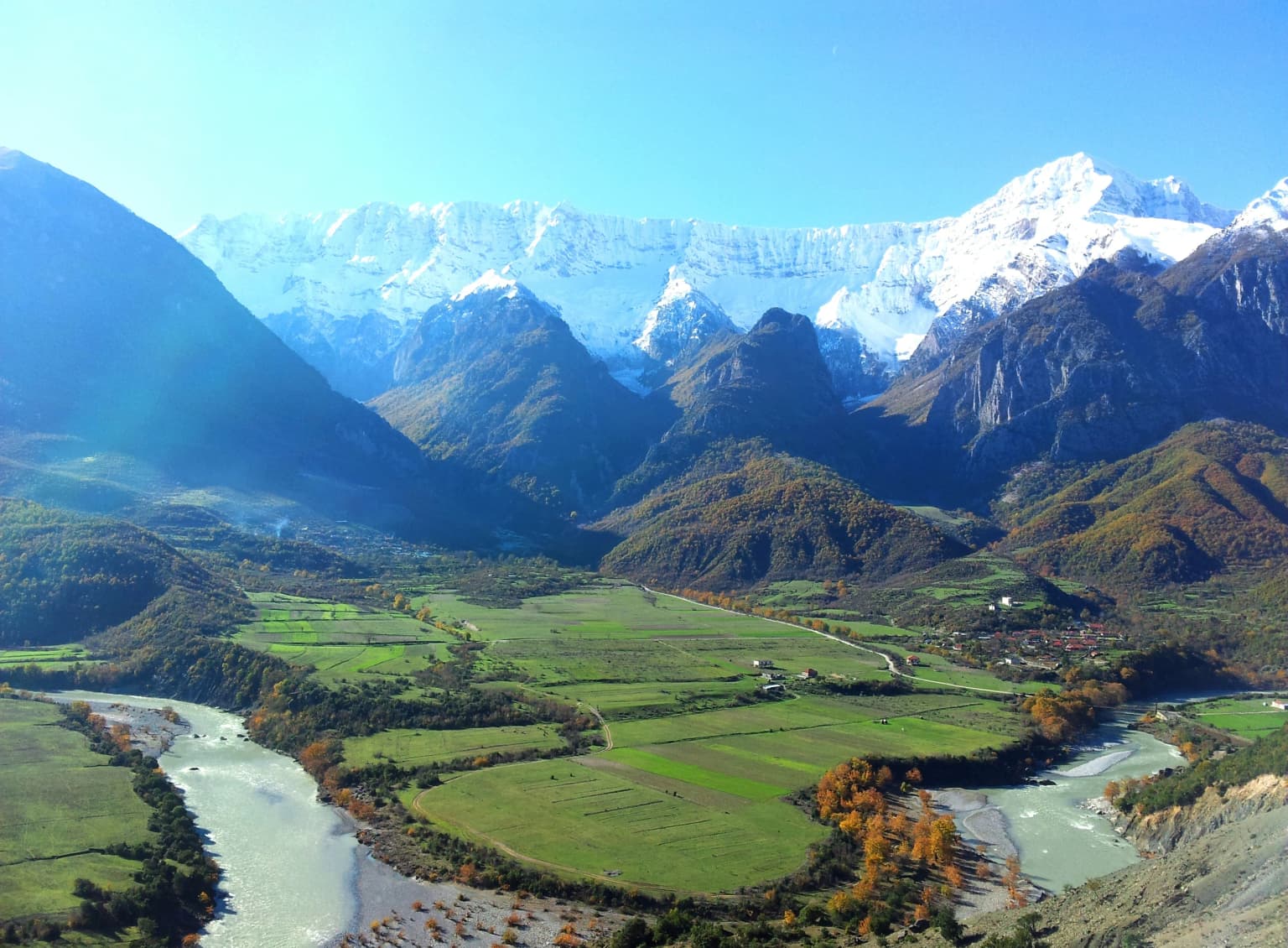 Wide landscape featuring the Vjosa River winding through green valleys with snow-capped mountains in the background under a clear blue sky.
