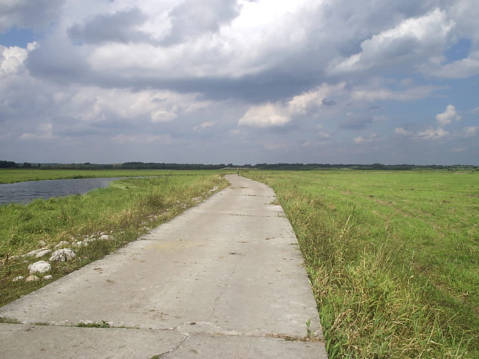 Concrete path running through grassy fields with a body of water on the left and cloudy sky above