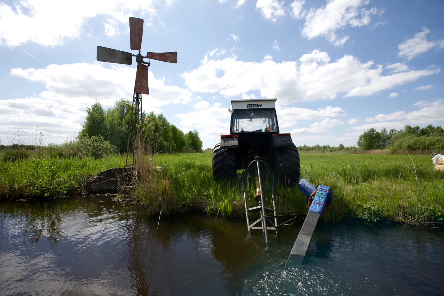 A water pump, tractor, and windmill situated by a waterway in a grassy wetland under a partly cloudy sky.