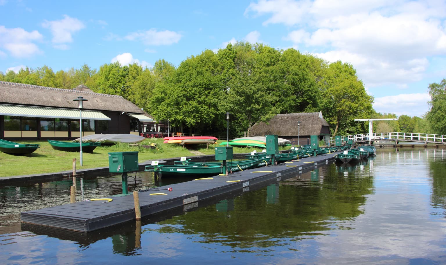 Floating dock with green boats, grassy shore, and thatched-roof buildings on calm water